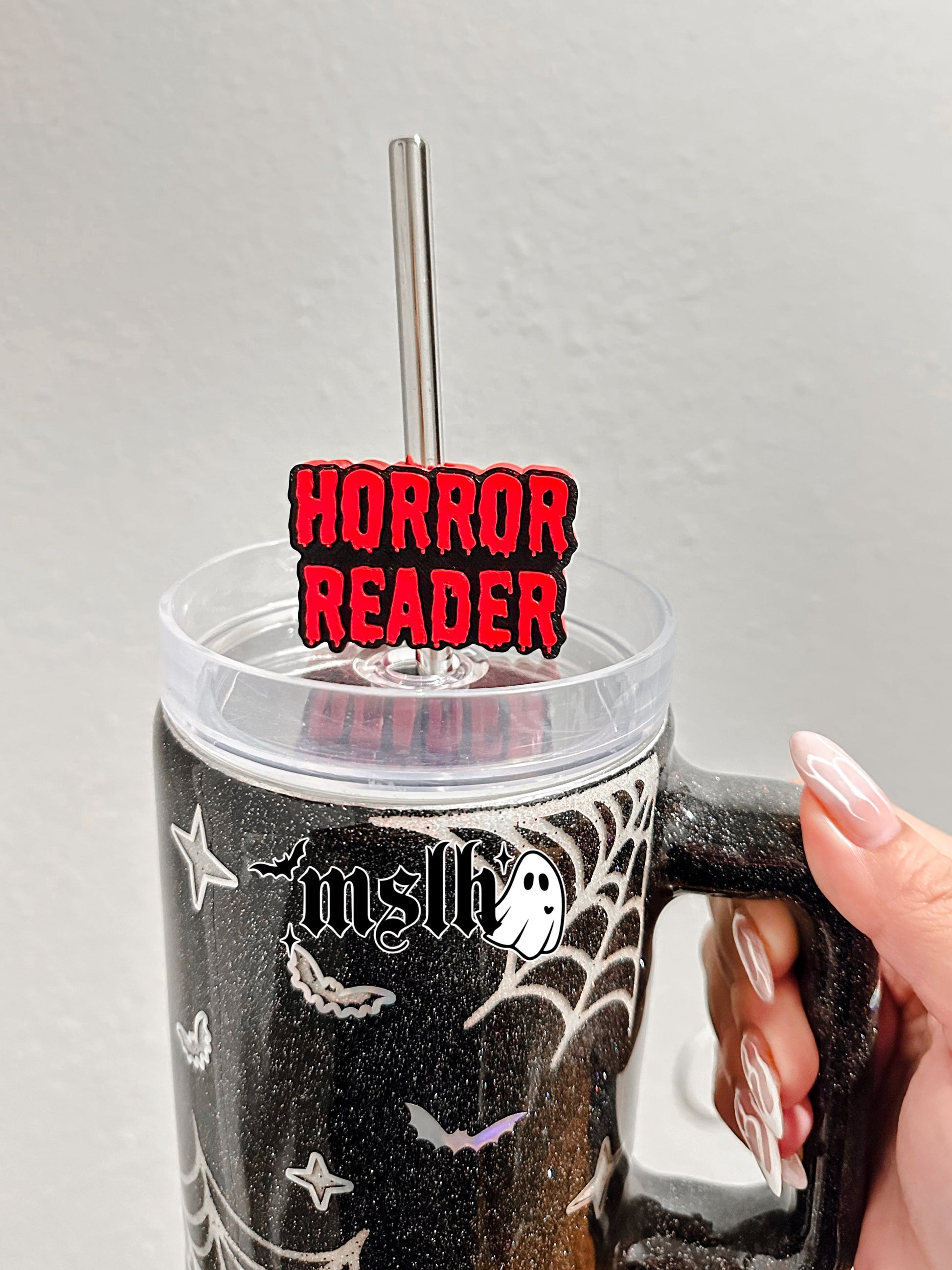 A hand holds a black glitter handled tumbler with spiderweb and bat graphics, a clear lid, and a metal straw. A red and black 'HORROR READER' sign is attached to the straw.