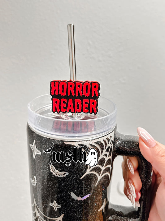A hand holds a black glitter handled tumbler with spiderweb and bat graphics, a clear lid, and a metal straw. A red and black 'HORROR READER' sign is attached to the straw.